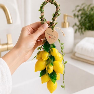 May include: A bunch of yellow lemon soaps with green leaves and a woven handle. The lemons are held by a hand. A heart-shaped tag reads "Handmade with love."
