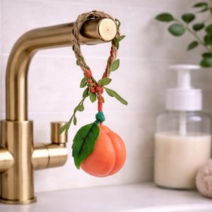 Może przedstawiać: Mydło w kształcie brzoskwini wisi na złotym kranie. Mydło jest pomarańczowe z dołączonym zielonym liściem. Jest zawieszone na sznurku ozdobionym zielonymi liśćmi i czerwonymi akcentami. Biała umywalka i dozownik mydła są w tle.