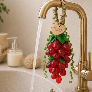 Op de afbeelding: Decoratieve zeep in de vorm van een tros rode druiven, hangend aan een messing kraan. De zeep heeft groene bladeren en een klein houten hartlabel met de tekst "Handmade with love". Water stroomt uit de kraan in een wastafel.
