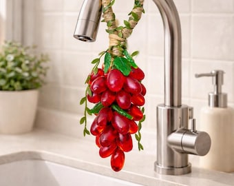 May include: A decorative kitchen faucet charm featuring a cluster of red grapes with green leaves. The grapes are attached to a twine and vine-wrapped loop, hanging from a stainless steel faucet. The charm adds a touch of whimsy to the kitchen.