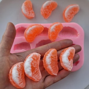 May include: Orange and white citrus fruit slices, possibly soap or candy, are displayed. A pink silicone mold with matching shapes is visible. The slices are held in a hand, with others arranged on a surface.