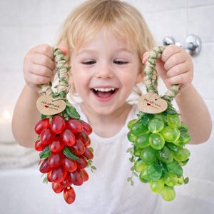Può includere: Un bambino tiene in mano due grappoli d'uva decorativi. Uno è rosso, l'altro verde. Ogni grappolo ha un manico intrecciato e un'etichetta con la scritta "Handmade with love".