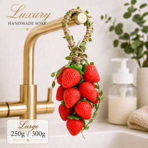 Puede incluir: Jabón artesanal con forma de racimo de fresas, colgado de un grifo. El jabón rojo tiene hojas verdes y lleva la inscripción "Luxury Handmade Soap". Peso: 250g / 300g.