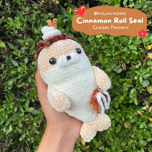 Puede incluir: Un juguete de peluche de foca de rollo de canela tejido a crochet con un patrón de piel marrón y blanco. El juguete sostiene un rollo de canela marrón y blanco en su aleta. El juguete está siendo sostenido en la mano de una persona. El texto "Cinnamon Roll Seal Crochet Pattern!" se muestra sobre el juguete.
