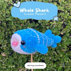 May include: A handmade blue and white crochet whale shark toy with black eyes and a pink mouth. The toy is mounted on a wooden stick and is set against a green leafy background. Text reads "Whale Shark Crochet Pattern!"