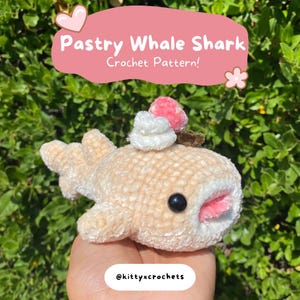 May include: A crocheted whale shark plush toy with a pink and white frosting topping. The toy is light brown with a pink mouth and black eyes. The text "Pastry Whale Shark Crochet Pattern!" is written above the toy.