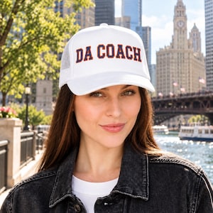 May include: A white trucker hat featuring a mesh back and a structured front panel. The text DA COACH is printed in a bold, collegiate-style font with dark blue lettering and a bright orange outline centered on the front of the cap.