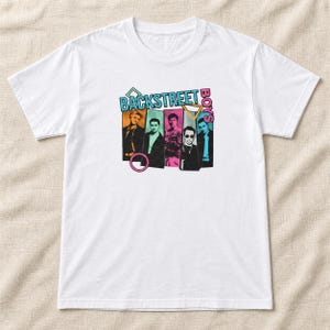 May include: A white short-sleeve t-shirt featuring a graphic print of the Backstreet Boys. The design includes five individual portraits of the band members in colorful rectangular frames, with the text BACKSTREET BOYS and geometric shapes.