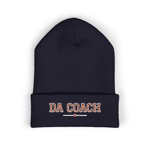 May include: A navy blue knit beanie with a cuffed brim. The front of the cuff features the text DA COACH embroidered in orange with a white outline, positioned above a horizontal white line with a small orange star in the center.