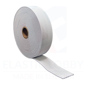May include: A roll of white elastic band, approximately 1 inch wide, is shown on a white background. The elastic is wound around a cardboard core. The elastic is made in the USA.