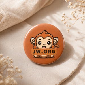 May include: A round, brown button featuring a cartoon monkey holding a sign that reads "JW.ORG". The monkey has large eyes and a smiling expression. The button is set against a neutral background.
