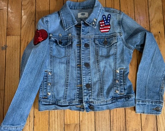 May include: A light blue denim jacket with silver studs, a red heart patch, and a peace sign patch with an American flag design.