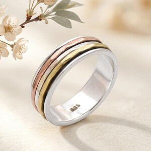May include: A silver ring featuring three bands in rose gold, gold, and silver. The ring has a hammered texture on the bands and the inscription "aas" is visible. The ring is displayed with a sprig of white flowers.