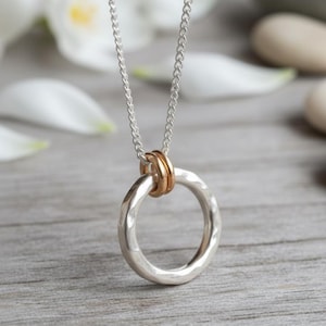 Puede incluir: Un collar plateado con un colgante en forma de círculo con textura martillada. Dos anillos dorados están unidos a la parte superior del círculo. La cadena del collar es plateada y descansa sobre una superficie de madera.