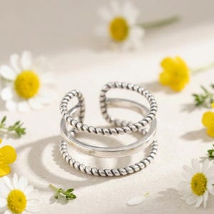 Può includere: Un anello color argento con un design a tre fasce. Le fasce superiore e inferiore hanno una trama a corda ritorta, mentre la fascia centrale è liscia. L'anello è aperto nella parte superiore. Margherite bianche e fiori gialli circondano l'anello.