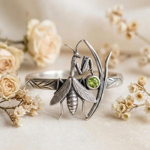 Puede incluir: Un anillo de plata con una escultura detallada de una mantis religiosa posada sobre un tallo curvo. La banda tiene un patrón de hojas grabado y una pequeña piedra preciosa de peridoto verde, redonda y facetada, está engastada junto a la mantis.