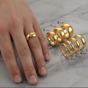 May include: A collection of gold rings of varying widths, displayed on a marble surface. The rings range from 2 mm to 8 mm in width. One ring is worn on a finger. The rings have a polished, reflective surface.