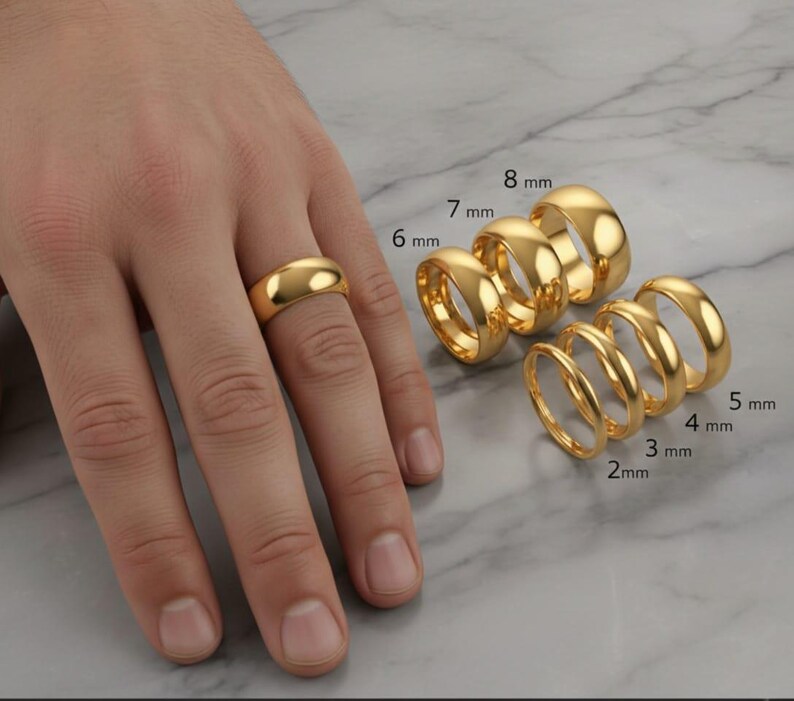 May include: A collection of gold rings of varying widths, displayed on a marble surface. The rings range from 2 mm to 8 mm in width. One ring is worn on a finger. The rings have a polished, reflective surface.