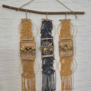 May include: Three macrame wall hangings with a natural wood branch at the top. The hangings are made with a combination of natural and black cord and feature dried flowers and leaves.