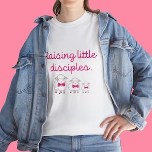 Könnte beinhalten: Weißes T-Shirt mit dem pinkfarbenen Text "Raising little disciples." und drei Cartoon-Schafen mit rosa Fliegen. Die Person trägt eine blaue Jeansjacke.