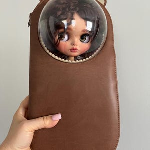 May include: A brown, elongated, bear-shaped carrying case with a clear, spherical, bubble-like window at the top. Inside the bubble window, a doll with dark, curly hair and large, expressive eyes is visible. The case features small, rounded ears.