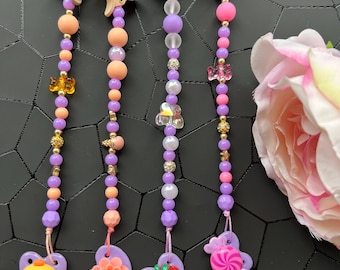 Puede incluir: Cuatro pinzas para el pelo con cuentas y abalorios decorativos. Cada pinza tiene un diseño diferente: un pollito amarillo, una flor, una fresa y un caramelo en espiral. Las cuentas son moradas, rosas y doradas, con una pinza a juego en la parte superior.