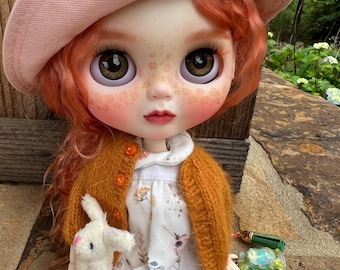 Può includere: Una bambola con capelli arancioni ondulati, lentiggini e grandi occhi marroni, che indossa un cappello da pescatore rosa, un cardigan in maglia giallo senape con bottoni arancioni e un vestito bianco con stampa botanica, tenendo un piccolo coniglio di peluche.