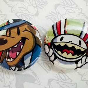Op de afbeelding: Twee ronde knopen met cartoonfiguren. De eerste knoop heeft een bruine hond met een blauwe hoed en een wit-blauw gepatroneerd shirt. De tweede knoop heeft een wit cartoonfiguur met een rood-wit gestreepte hoed en een groen-witte achtergrond.