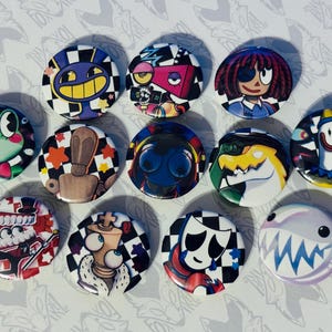 Op de afbeelding: Een verzameling van elf ronde, kleurrijke knoopspelden met verschillende cartoonkarakterontwerpen. De knoopspelden hebben een verscheidenheid aan kleuren, waaronder blauw, groen, rood en geel, met geblokte en patroonachtergronden. De ontwerpen omvatten een kikker, een robot en een haai.