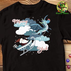 以下が含まれることがあります： サンタクロースがそりで飛んでいる青いドラゴンが描かれた黒いTシャツ。Tシャツには「メリーユール」の文字がプリントされています。