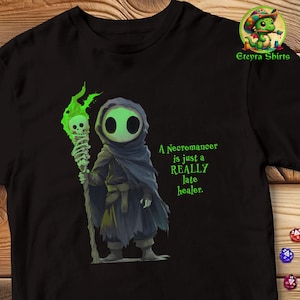 Può includere: Maglietta nera con un'illustrazione verde e bianca di una figura incappucciata che tiene uno scettro con un teschio in cima. Il testo "A Necromancer is just a REALLY late healer." è stampato in verde sulla maglietta.