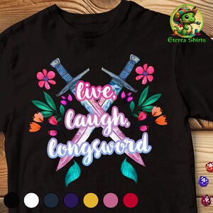 May include: Black t-shirt featuring a graphic of two crossed swords with pink and blue blades. The swords are surrounded by pink, purple, and green flowers. The text "live, laugh, longsword" is written in a purple font.