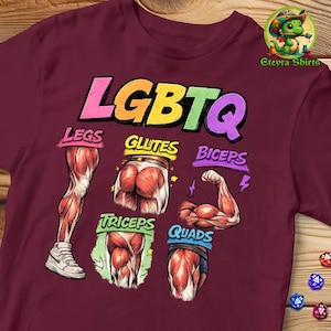 May include: A maroon t-shirt featuring a graphic with the text LGBTQ in rainbow colours. Below, anatomical illustrations of muscles are labelled Legs, Glutes, Biceps, Triceps, and Quads in colourful text. A small green dragon logo is in the top right corner.