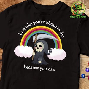 Könnte beinhalten: Schwarzes T-Shirt mit einer weißen Grafik eines Cartoon-Sensenmanns, der eine Sense hält und vor einem Regenbogen steht. Der Text lautet "Live like you're about to die because you are."
