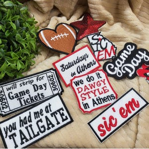Puede incluir: Una colección de parches para planchar que incluyen un balón de fútbol, un corazón, una estrella y varios textos como "Game Day", "Saturdays in Athens", "We do it Dawgy Style in Athens" y "sic'em".
