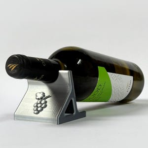 Può includere: Un portabottiglie per vino color argento con un design a grappolo d'uva. Una bottiglia di vino verde è posizionata orizzontalmente nel supporto. La bottiglia ha un tappo nero e un'etichetta verde e bianca. Il supporto è in materiale chiaro.