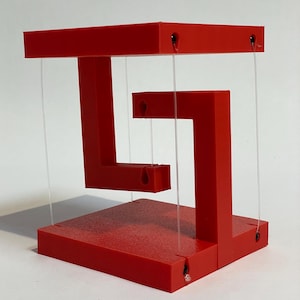 May include: A red geometric sculpture with a unique design. The sculpture appears to defy gravity, with its components suspended by clear strings. The structure is made of solid red material, creating a striking visual contrast against the white background.