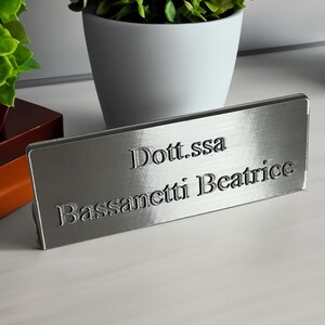 Puede incluir: Una placa de identificación plateada con el texto grabado "Dott.ssa Bassanetti Beatrice" se muestra sobre una superficie clara. La placa es rectangular y tiene un acabado de metal cepillado. Una maceta blanca con una planta verde está en el fondo.