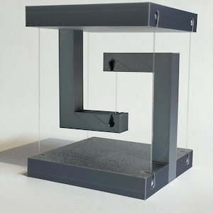May include: A gray, geometric sculpture with a floating design. The sculpture features a top and bottom square connected by vertical supports, with a suspended, angled section. Clear strings hold the floating elements in place.