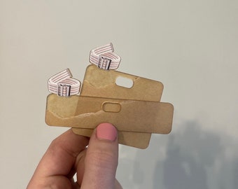 May include: Two white and pink striped elastic bands with silver buckles are stacked on top of a brown cardboard tag with a rectangular cutout.