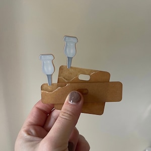 May include: A close-up of a brown paper card with two white plastic push pins. The push pins are attached to the card with a small amount of adhesive.