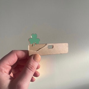 May include: A St. Patrick's Day themed hair clip featuring a light green shamrock on a rectangular, tan-colored base. The base has a small hole and a torn edge. The clip is held against a plain, light background.