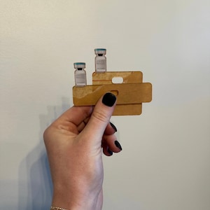 May include: A wooden keychain with two small glass vials labelled "DANNER'S" on a shelf. The keychain is held in a hand with black nail polish.