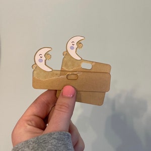 Puede incluir: Dos fundas de teléfono de cartón marrón con un diseño de media luna blanca. Cada luna tiene un pequeño círculo morado y una pequeña estrella amarilla.