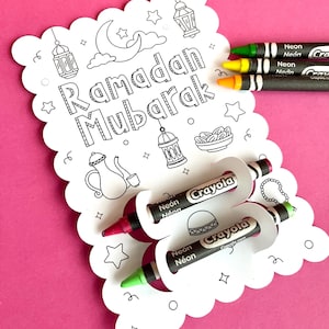 Puede incluir: Una tarjeta blanca con una luna creciente, estrellas y linternas. La tarjeta dice "Ramadan Mubarak" en letras negras. Hay cuatro crayones Crayola de neón en la tarjeta.
