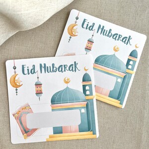 K&ouml;nnte beinhalten: Zwei wei&szlig;e Karten mit dem Text "Eid Mubarak" in schwarzer Kalligraphie. Jede Karte zeigt eine farbenfrohe Illustration einer Moschee mit einem Halbmond und einem Stern, einem Gebetsteppich und einer Laterne.