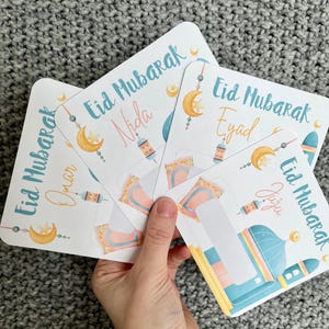 K&ouml;nnte beinhalten: Vier bunte Karten mit dem Text "Eid Mubarak" und verschiedenen Namen auf jeder Karte. Die Karten zeigen einen Halbmond, eine Moschee und eine Laterne.
