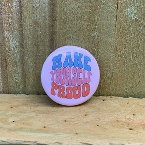 May include: A pink button with the text "MAKE YOURSELF PROUD" in blue, pink, and orange wavy letters.