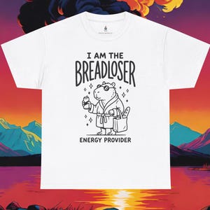 May include: A white t-shirt featuring a black line art graphic of a capybara wearing sunglasses and a bathrobe, holding a drink and a bag with a baguette. The text reads I AM THE BREADLOSER ENERGY PROVIDER in black lettering above and below the graphic.