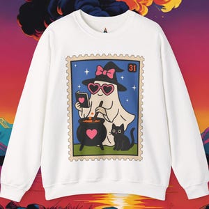 May include: White sweatshirt featuring a Halloween-themed graphic. The graphic depicts a ghost wearing a witch hat and heart-shaped glasses, stirring a cauldron with a black cat nearby. The graphic is framed like a postage stamp.